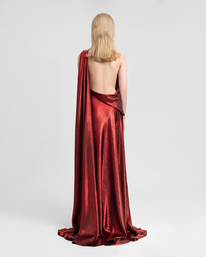 Back view of an asymmetrical lamé georgette gown featuring a dramatic open back and a one-sided cape that flows to the floor. The metallic red fabric shimmers under the light, enhancing the dress’s fluid, elegant silhouette.
