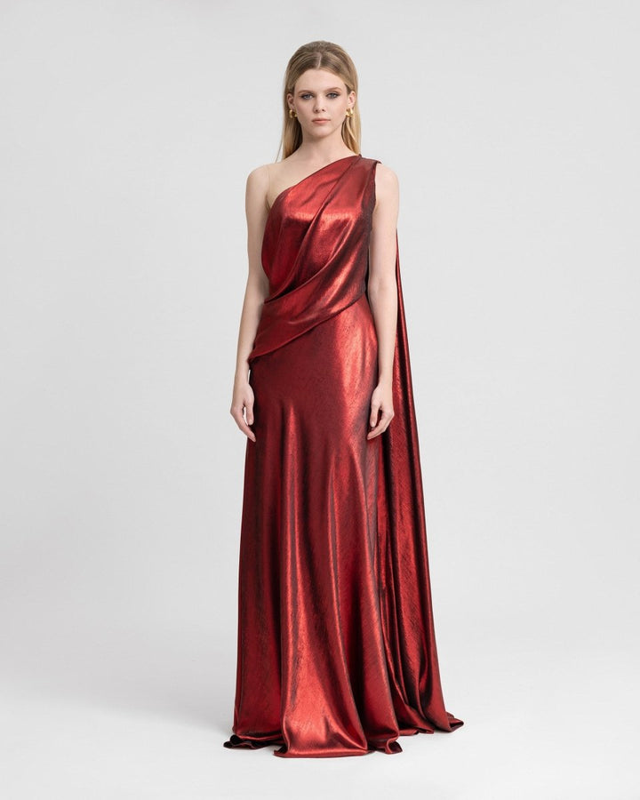 Model wearing an asymmetrical lamé georgette gown with a draped bodice and a one-sided flowing cape. The shimmering metallic finish and open back add a dramatic, elegant touch to this red-carpet-ready evening dress.