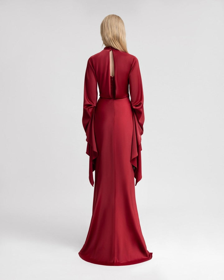 Back view of a woman wearing a deep red satin gown with a floor-length hem and a keyhole opening at the upper back. The dress features long, ruffled sleeves that drape elegantly down the sides, enhancing its dramatic and graceful silhouette.