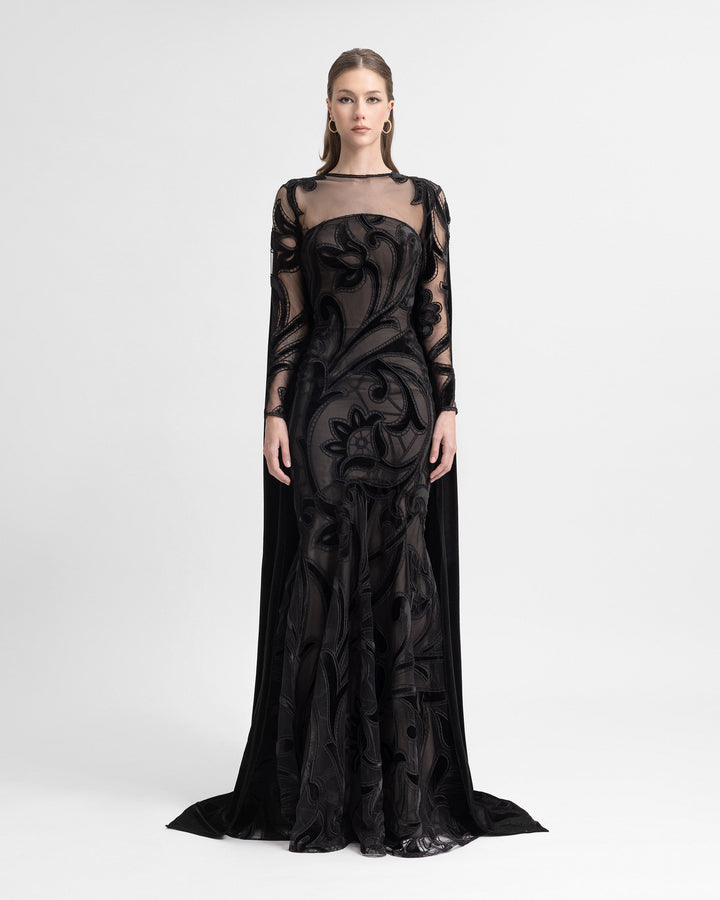 Woman wearing a strapless slim-cut black embroidered velvet dress with a sheer mesh cape overlay, featuring long sleeves and a floor-length hemline.