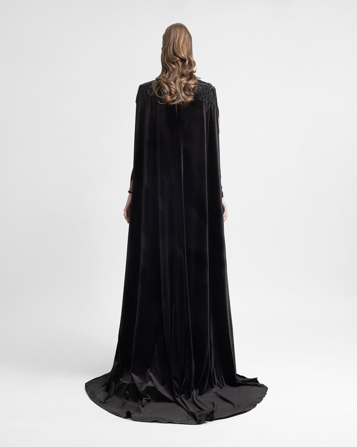 Back view of a woman in a black velvet gown with an attached floor-length cape, creating a dramatic, flowing silhouette.