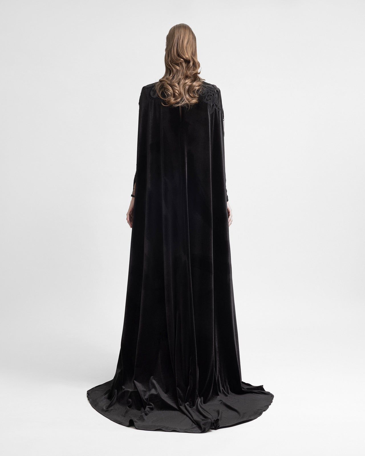 Back view of a woman in a black velvet gown with an attached floor-length cape, creating a dramatic, flowing silhouette.