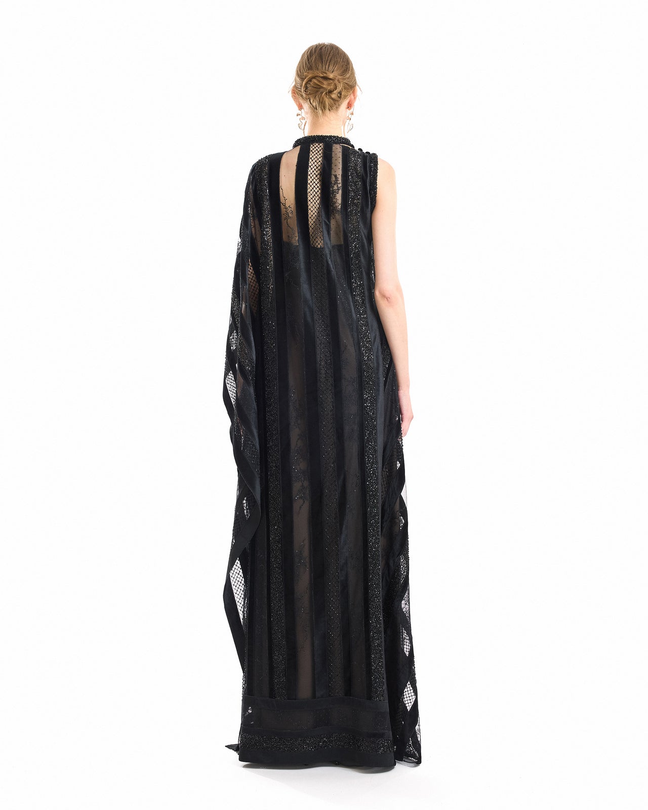 A woman viewed from the back wearing a sleeveless black maxi dress with sheer mesh panels, vertical lace-like stripes, and embroidered trim, photographed against a plain white studio background.