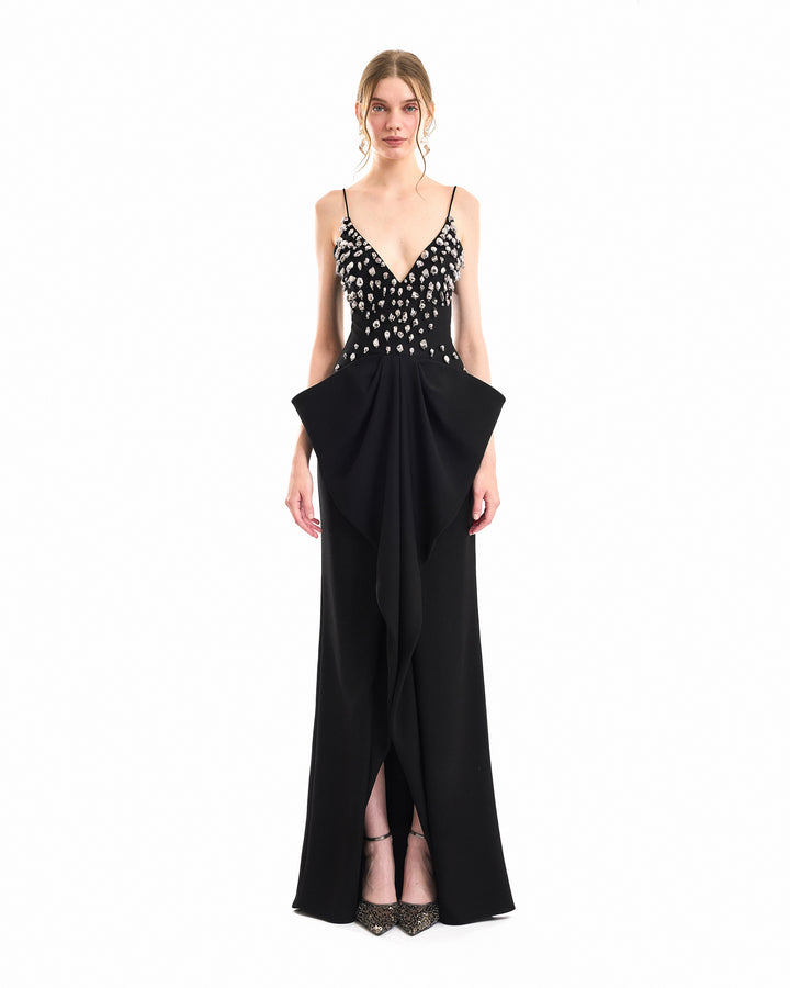 A model wearing a black floor-length gown with a beaded bodice, thin spaghetti straps, and a dramatic draped skirt detail, photographed against a solid white background.