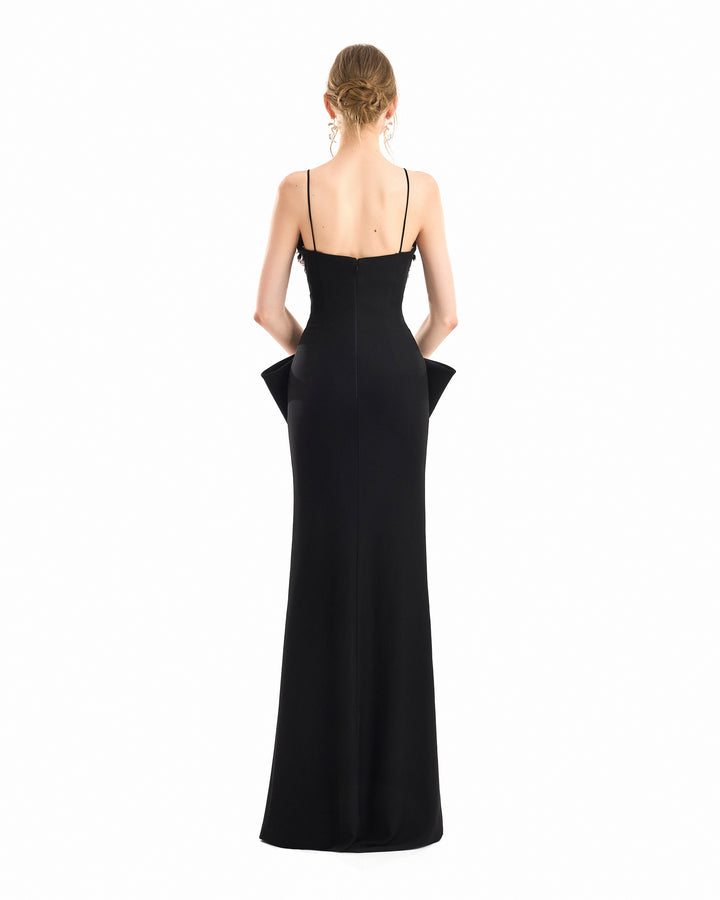 A woman viewed from the back wearing a floor-length black gown with thin spaghetti straps, a structured back, and dramatic flared ruffle details at the waist and hips, set against a plain white background.
