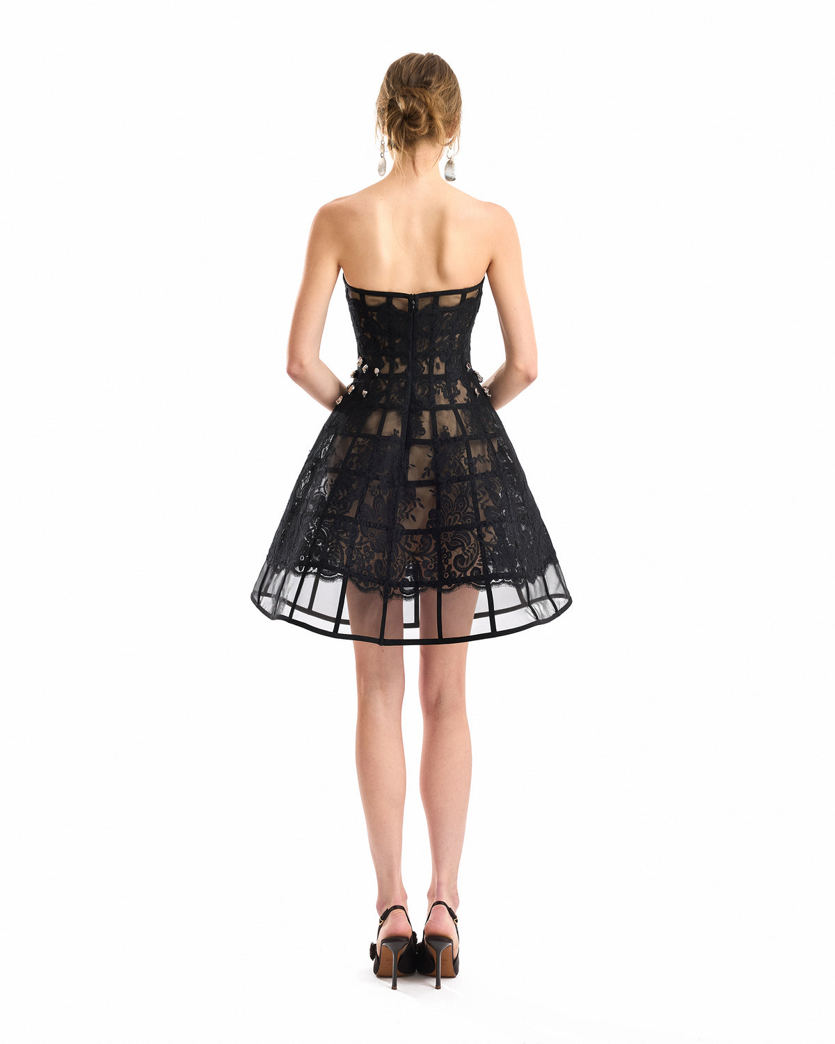 A woman viewed from the back wearing a strapless black lace mini dress with a fitted bodice and flared skirt, paired with black heels, on a plain white background.