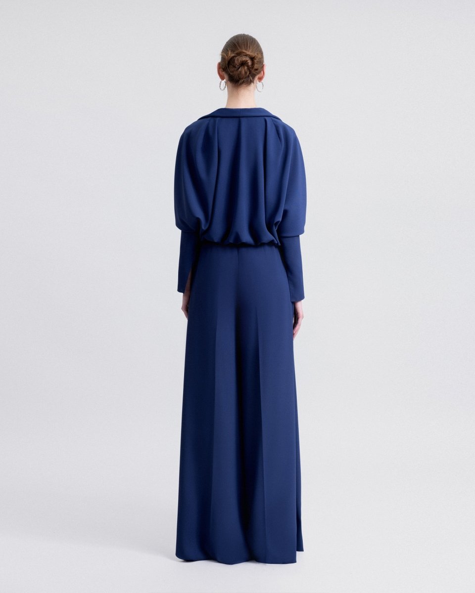 Back view of a model wearing a navy draped bolero with long sleeves over high-waisted straight-cut pants, showcasing the loose, elegant silhouette of the ensemble.