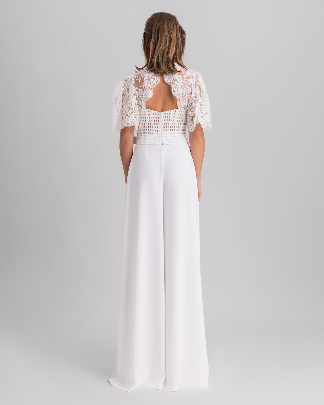The back of a white evening wear set featuring a lace top with flared sleeves and an open back,  paired with flared crepe pants.