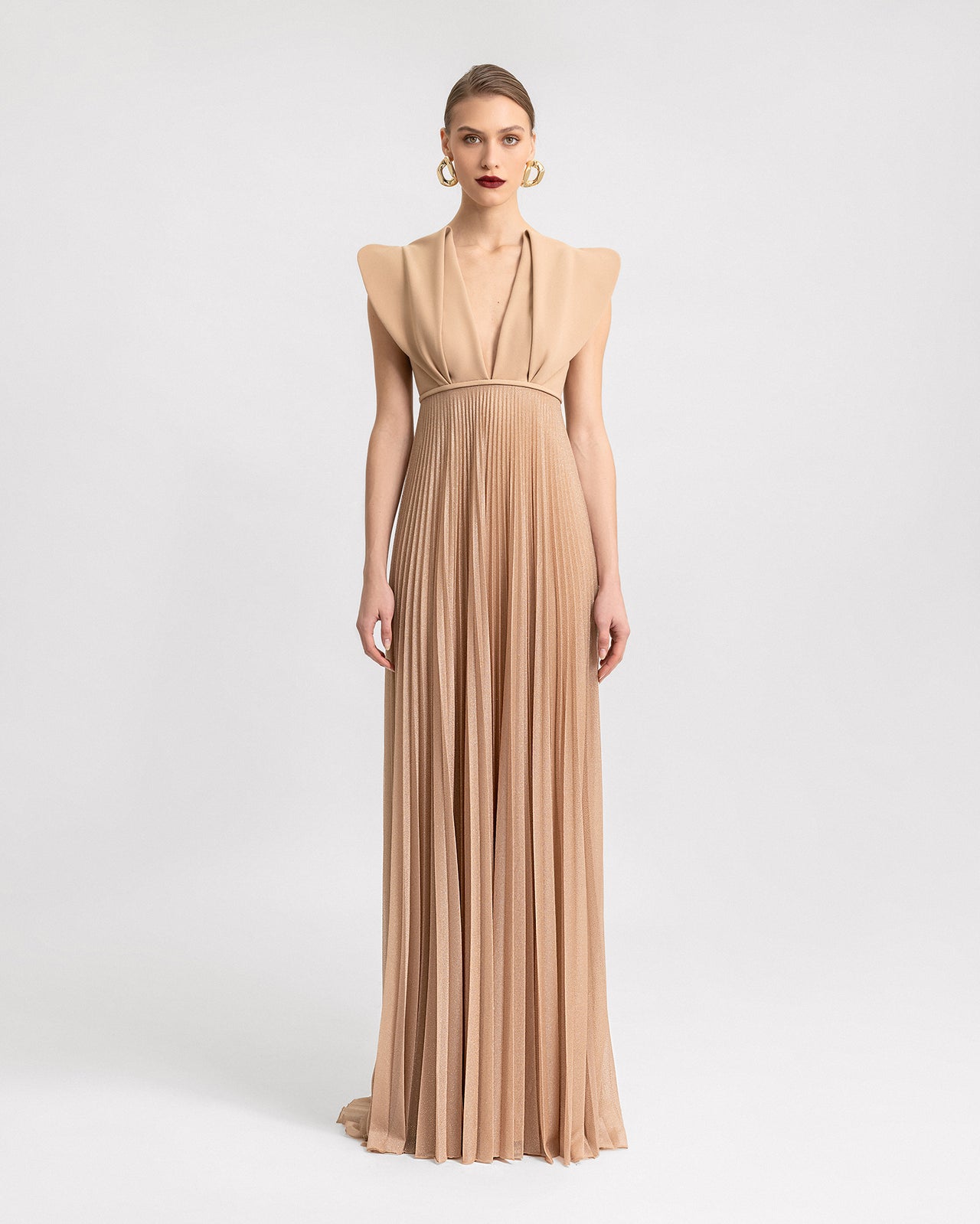 Video of a long beige dress with structured shoulders, a deep V-neckline, and a pleated shimmer skirt, showcasing the design’s movement and elegant silhouette from different angles.