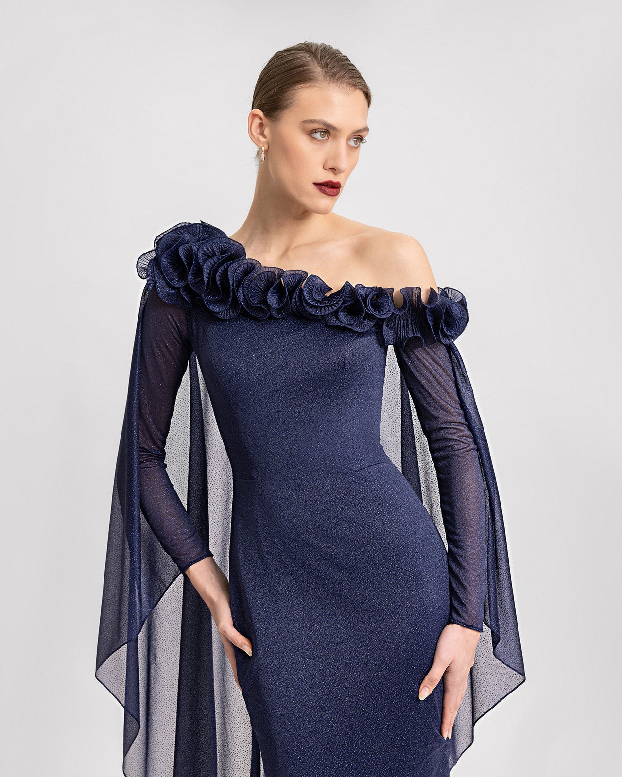 Video of a navy blue asymmetrical shimmer gown with a floral neckline, sheer sleeves, and a dramatic cape. The dress features a fitted silhouette and flows elegantly as the model moves.