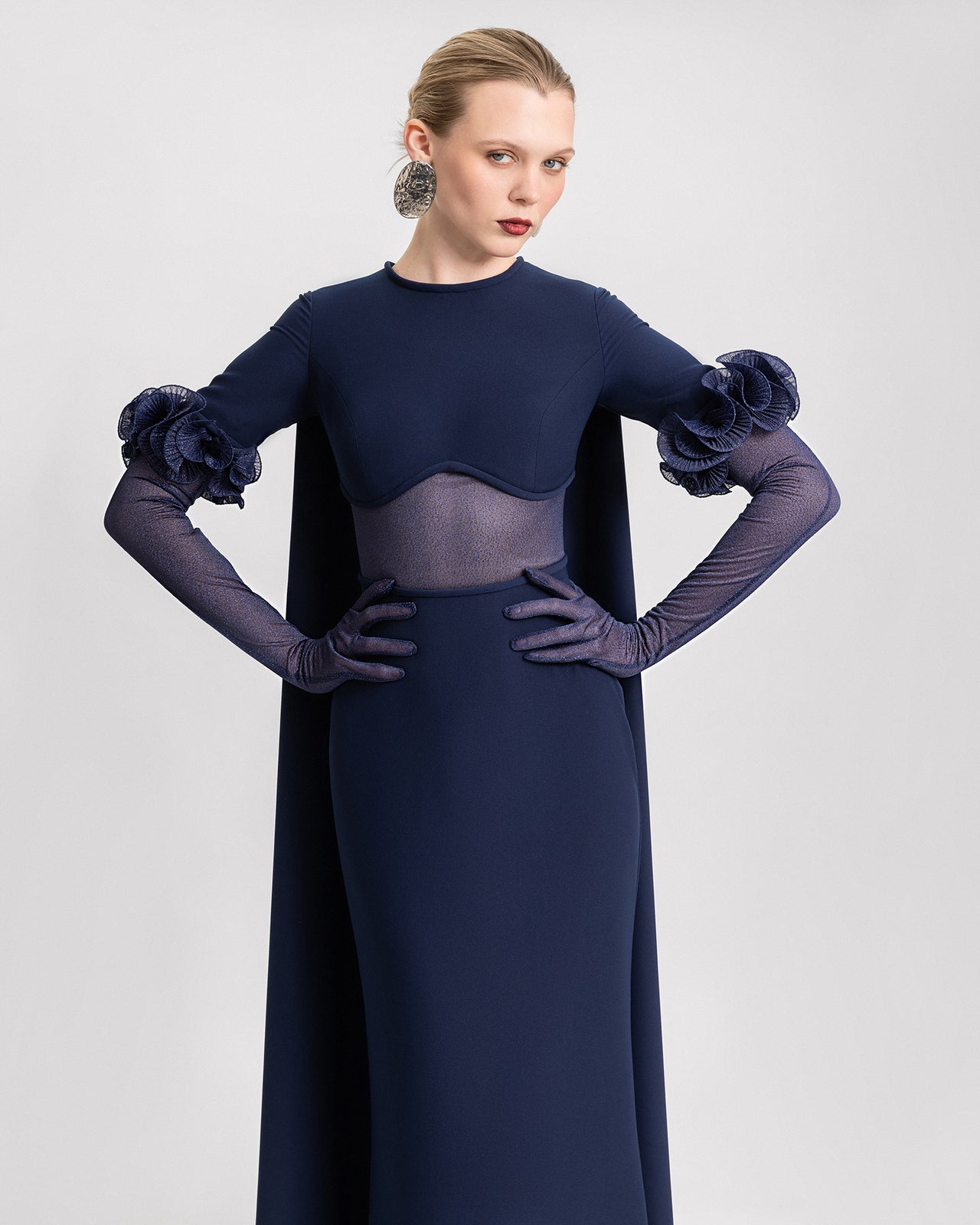 Video of a navy blue slim-fit gown with a sheer under-bust panel, detachable shimmer sleeves adorned with floral details, and a dramatic floor-length cape.

