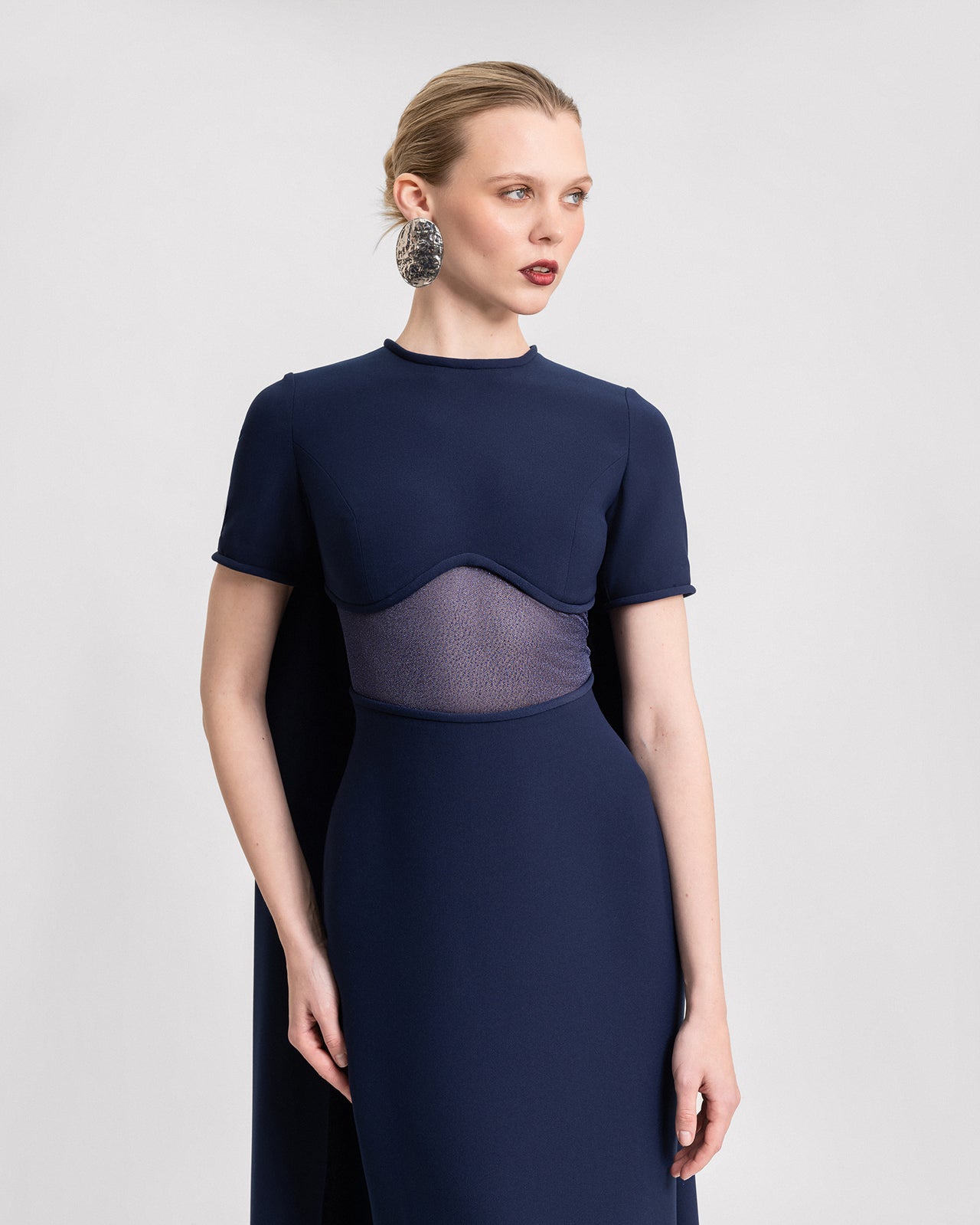 Video of a navy blue slim-fit gown with a sheer under-bust panel, short sleeves, and a dramatic floor-length cape. Shown without the detachable sleeves for a sleek, minimalist look.