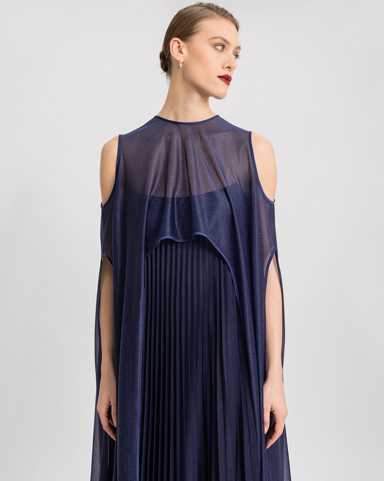 Video showcasing a navy blue strapless pleated gown paired with a long, sheer shimmer cape. The cape features elegant shoulder cut-outs and flows dramatically as the model walks, highlighting the dress’s fluid movement and luxurious texture.