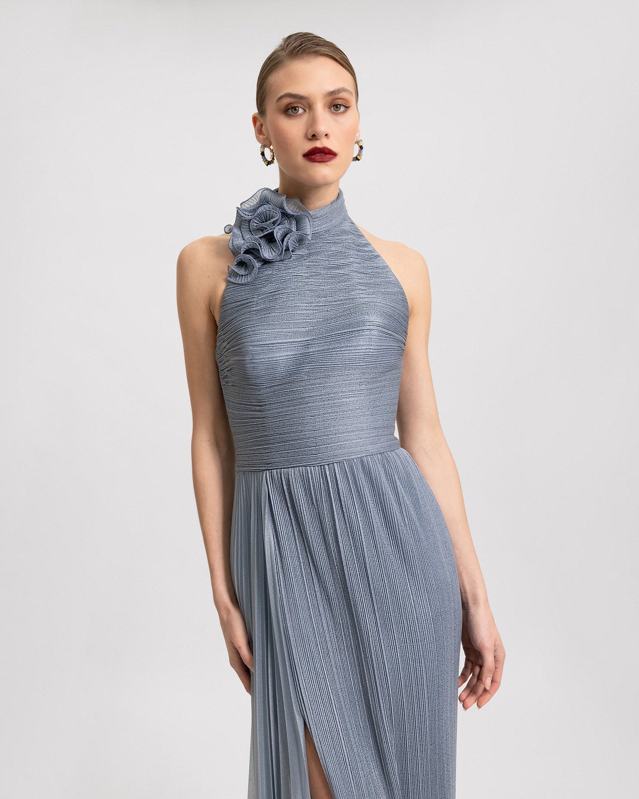 Video of a dusty blue shimmer dress with a high collar, floral neckline detail, pleated fabric, side slit, and open back, shown from multiple angles in motion.