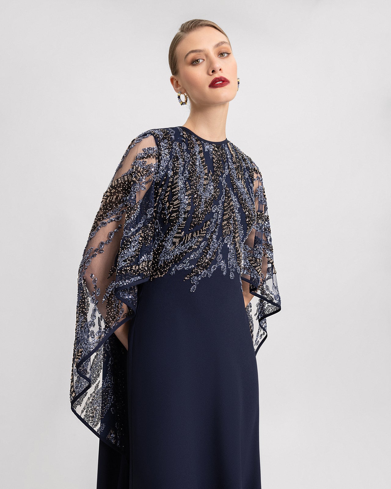 Video of a navy blue floor-length gown with sheer, beaded cape-like sleeves. The dress flows elegantly with every movement, showcasing the intricate embroidery on the upper bodice and sleeves.
