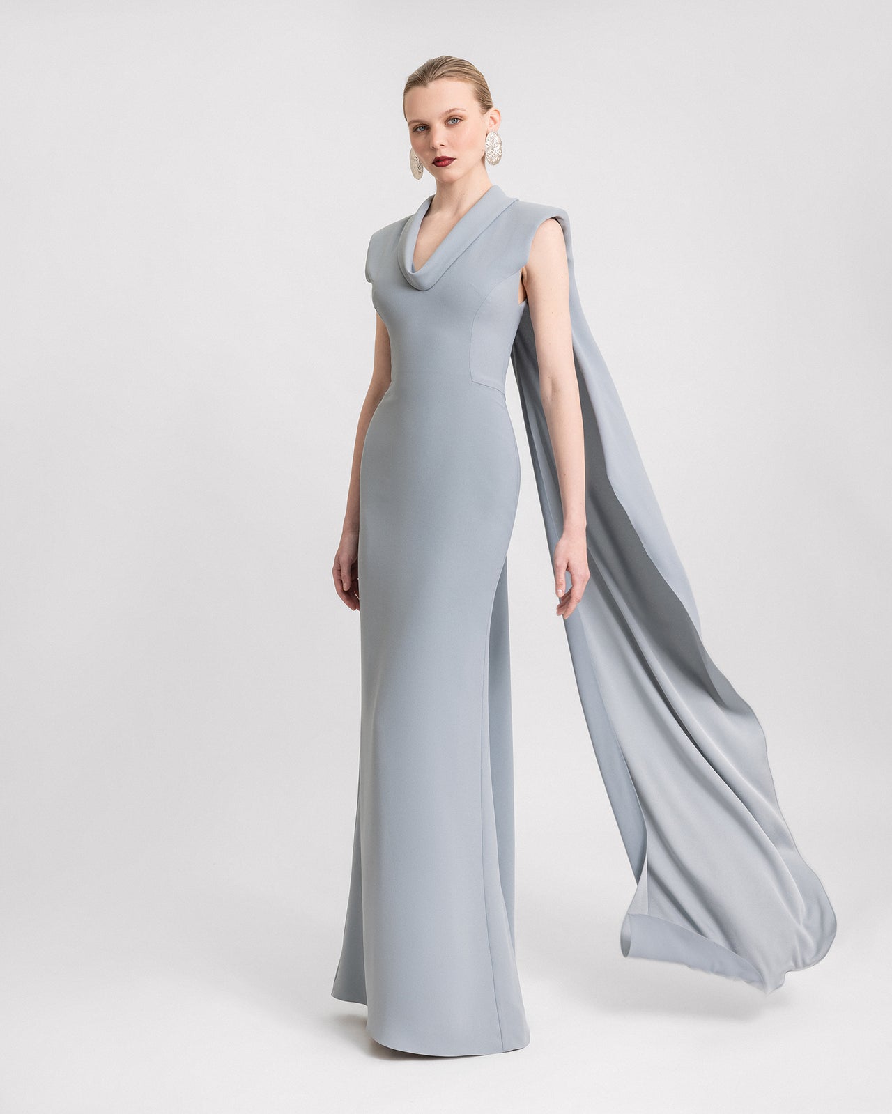 Video of a slim-cut teal blue dress with padded shoulders, a draped neckline, and an asymmetrical back cape, shown from multiple angles in motion to highlight the design and flow.