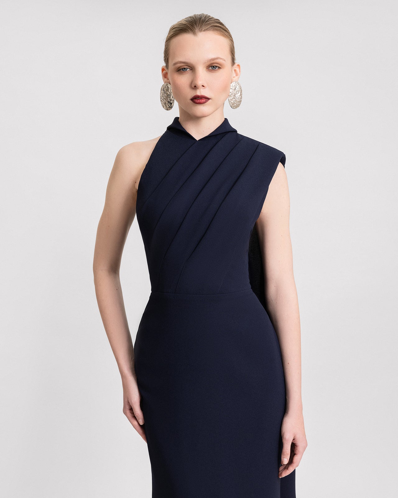 Video of a navy blue asymmetrical slim-fit gown with a padded shoulder, pleated bodice, and a long flowing cape. The dress moves elegantly with each step, showcasing its structured crepe fabric.