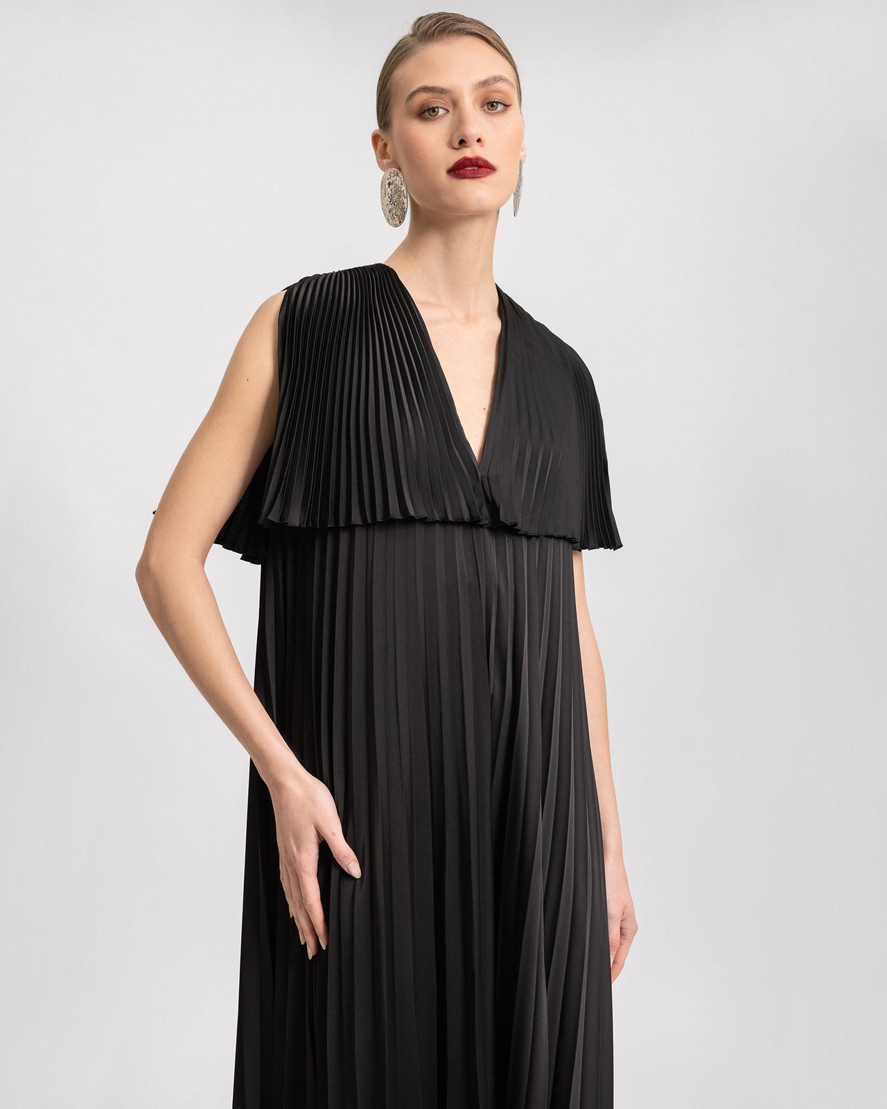 Video of a black plissé dress with a V-neckline, short sleeves, and shoulder cut-outs. The camera captures the flow and movement of the pleated satin fabric as the model walks, highlighting the dress's elegant, loose silhouette.