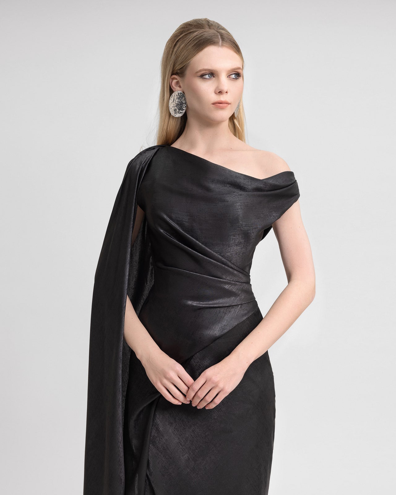 Video of a model wearing a black lamé georgette dress with an asymmetrical neckline and floor-length sleeve. The model moves to show off the fluid drape, sculpted fit, and elegant sheen of the fabric from all angles.