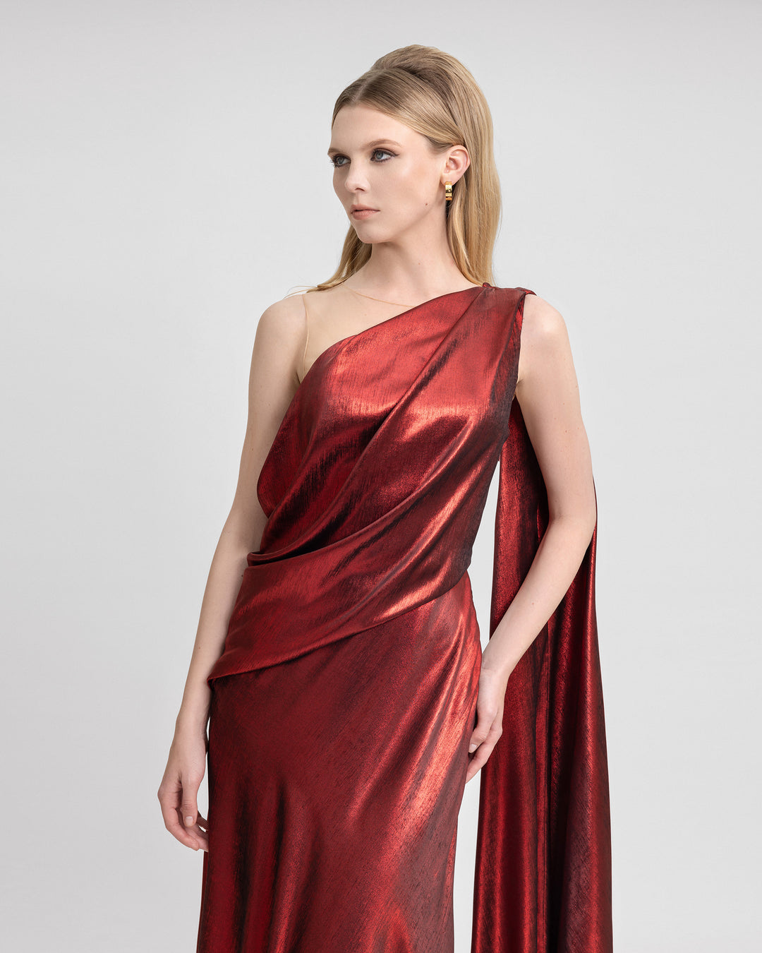 Video of a model wearing an asymmetrical red lamé georgette gown with a one-shoulder draped design, flowing cape, and open back. As the model moves, the metallic fabric shimmers and the cape trails elegantly, highlighting the dress’s fluid silhouette and glamorous finish.