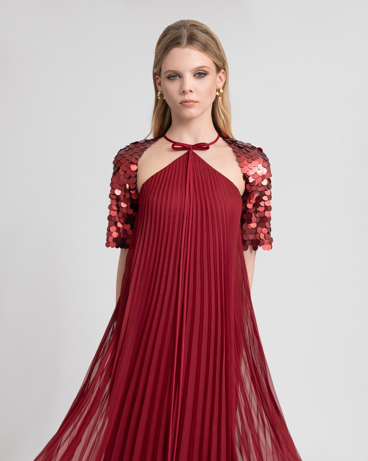 Video of a model wearing a halter-neck plissé chiffon gown with shimmering sequin bolero sleeves and a bow detail at the neckline. The dress sways gracefully as the model moves, showcasing the fluid pleats and eye-catching contrast between the soft fabric and glossy sequins—ideal for evening or red carpet looks.