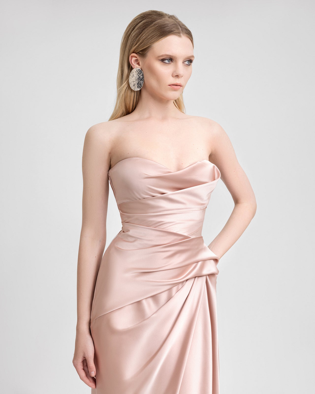 Video of a model wearing a blush pink strapless satin gown with asymmetrical draping along the waistline and a floor-length hem. As the model moves, the glossy satin catches the light, emphasizing the dress’s sculpted fit and soft, elegant flow.