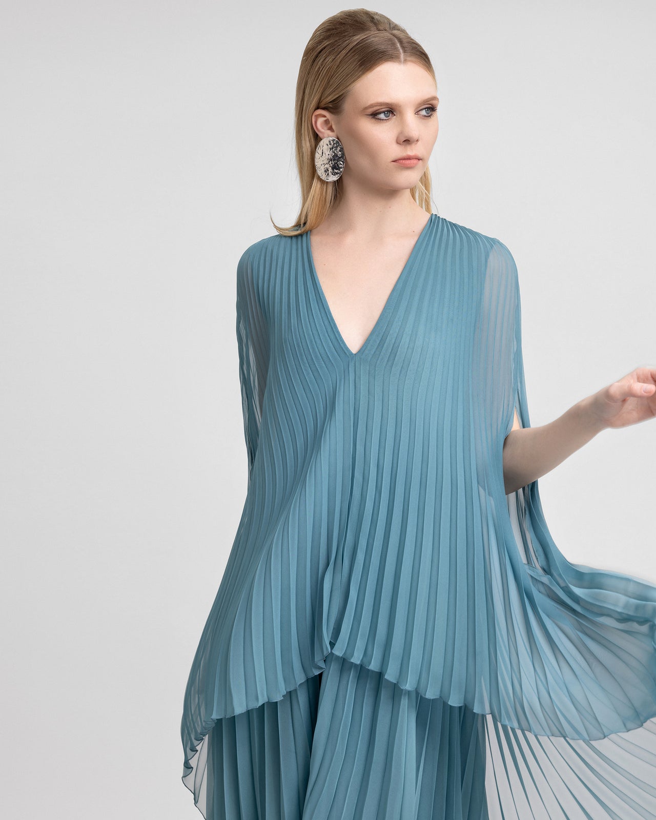 Video of a model wearing a soft blue chiffon plissé gown with a V-neckline and flowing cape-like sleeves. As the model moves, the pleated fabric sways gently, emphasizing the dress’s light, ethereal quality and graceful silhouette.