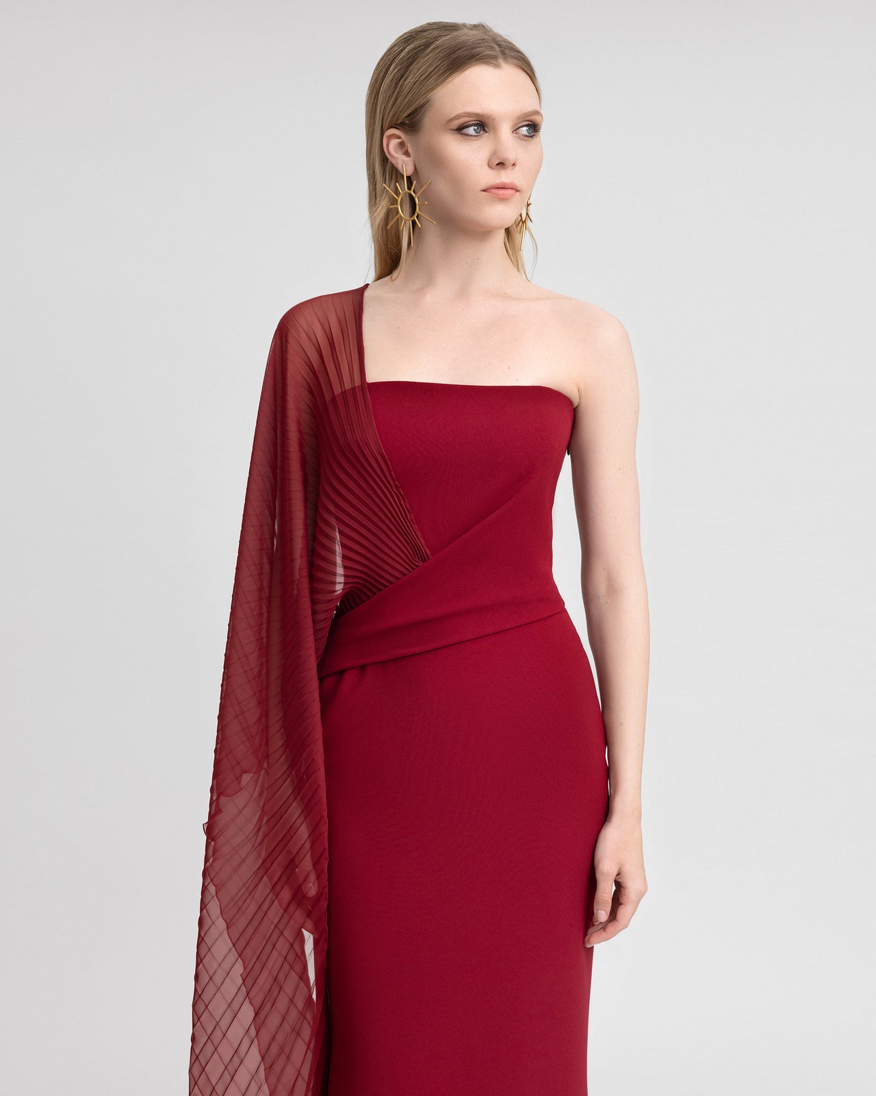 Video of a model showcasing a slim-cut red gown made of crepe and chiffon, featuring an asymmetrical draped waistline and a sheer, pleated floor-length sleeve. The model walks to reveal the fluidity of the chiffon and a subtle back slit, emphasizing the gown’s elegant movement and one-shoulder design.