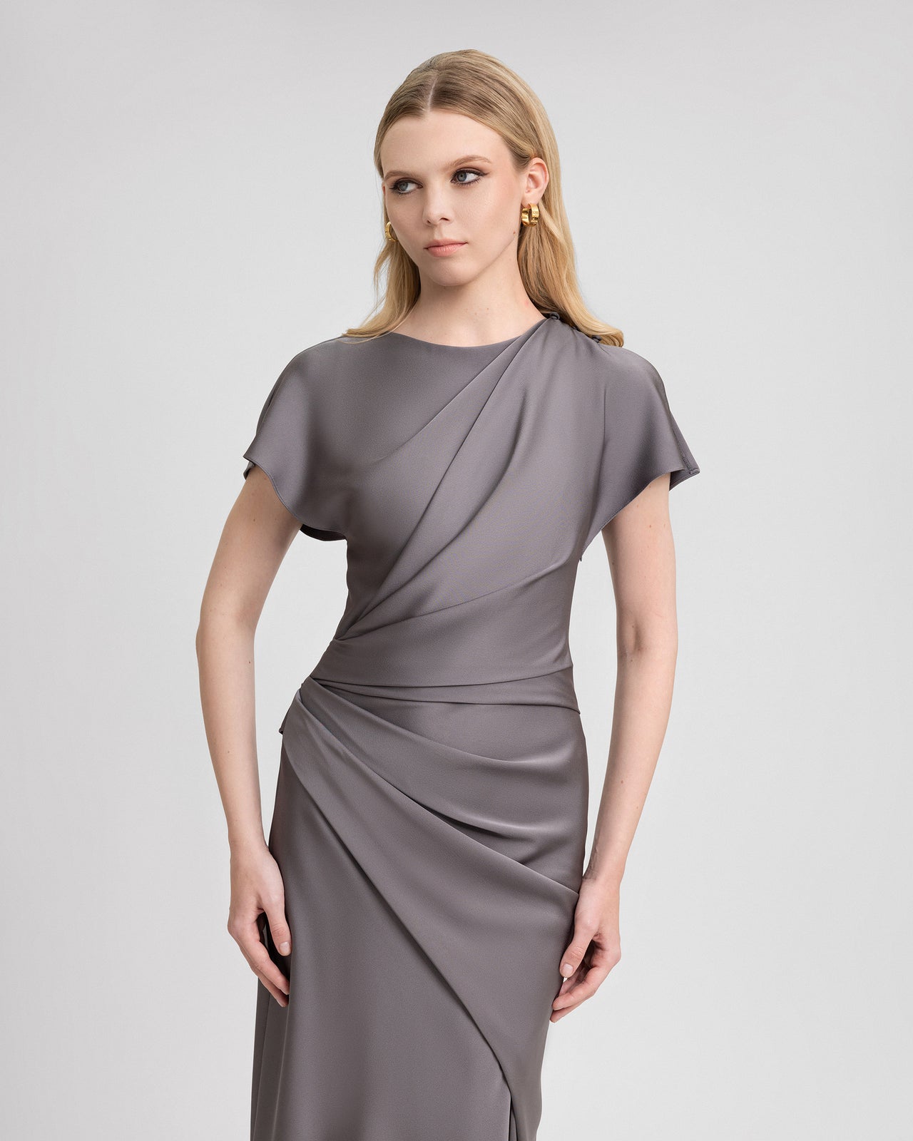 Video of a model wearing a smokey grey satin gown with asymmetrical draping, short sleeves, and a floor-length silhouette. As the model moves, the satin fabric catches the light, highlighting the fluid drape and elegant construction of this refined evening dress.