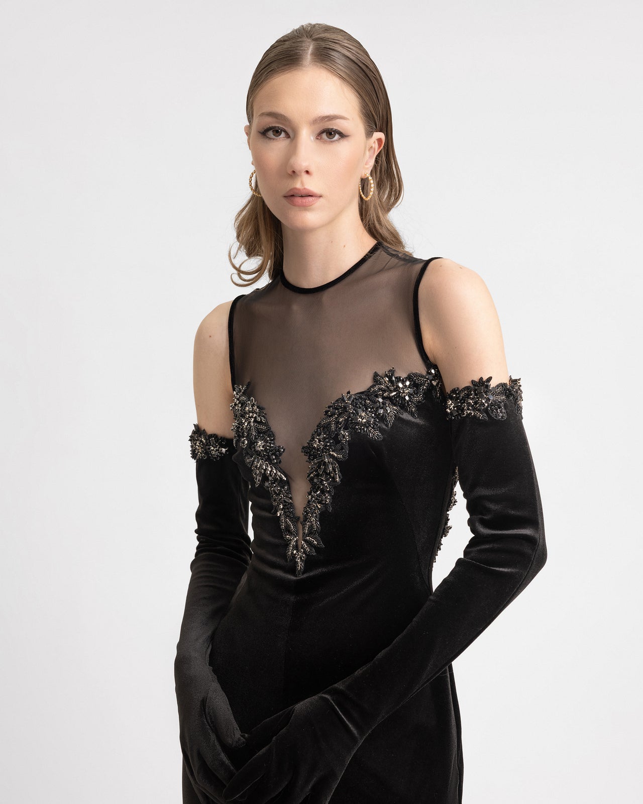 Video of a model wearing a black velvet gown with a sheer mesh bodice and detachable velvet gloves. The dress features a deep heart-shaped neckline with intricate beadwork and a fitted floor-length silhouette, shown in motion to highlight the elegant drape and glove detail.