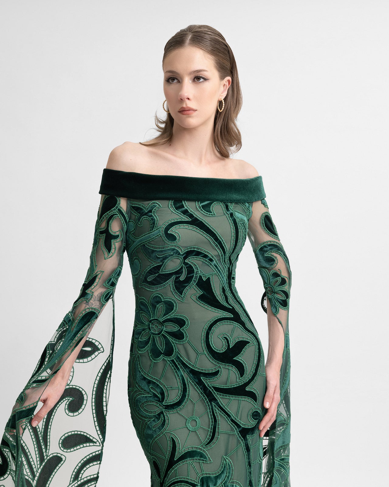 Video of a model wearing an off-the-shoulder emerald green gown with sheer floor-length sleeves, velvet floral embroidery, and a back slit, highlighting the dress’s elegant movement and intricate texture.