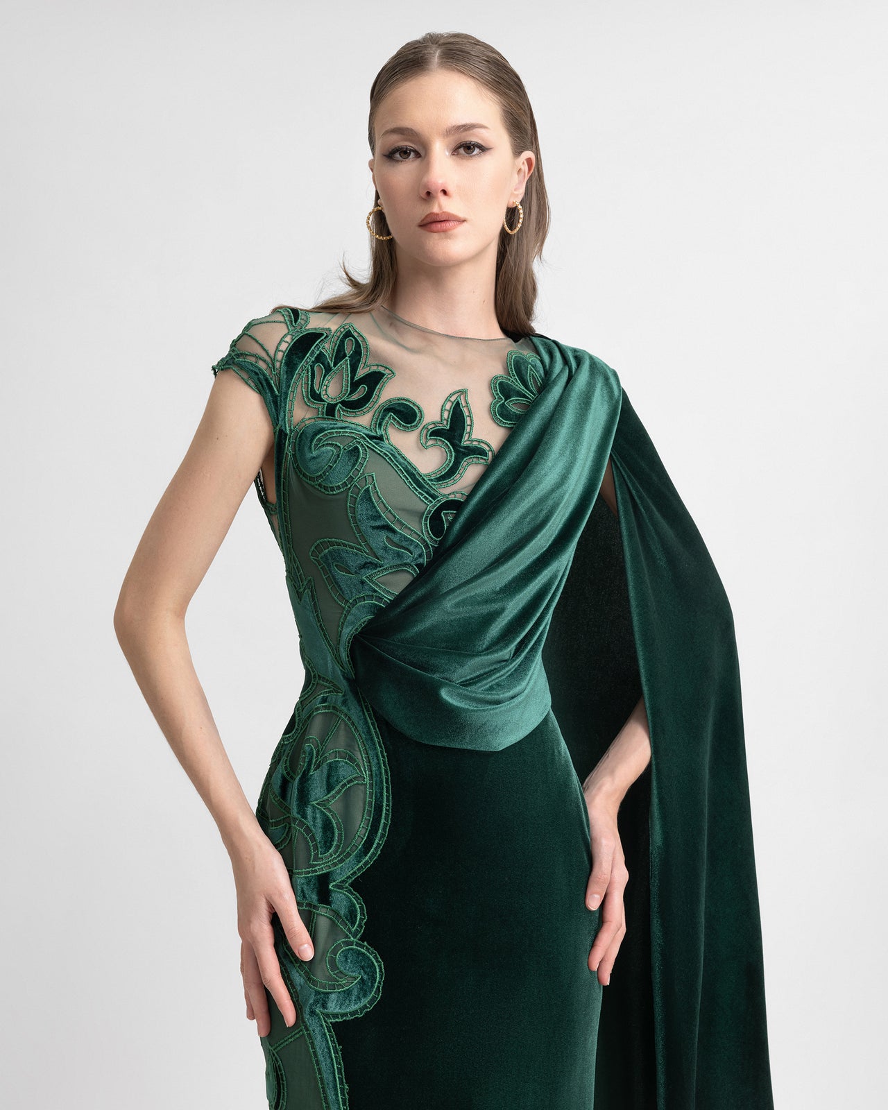 Video of a model wearing an emerald green asymmetrical velvet gown with floral embroidery, sheer bodice, and a one-sided draped cape, highlighting the dress’s flowing movement and intricate detailing.