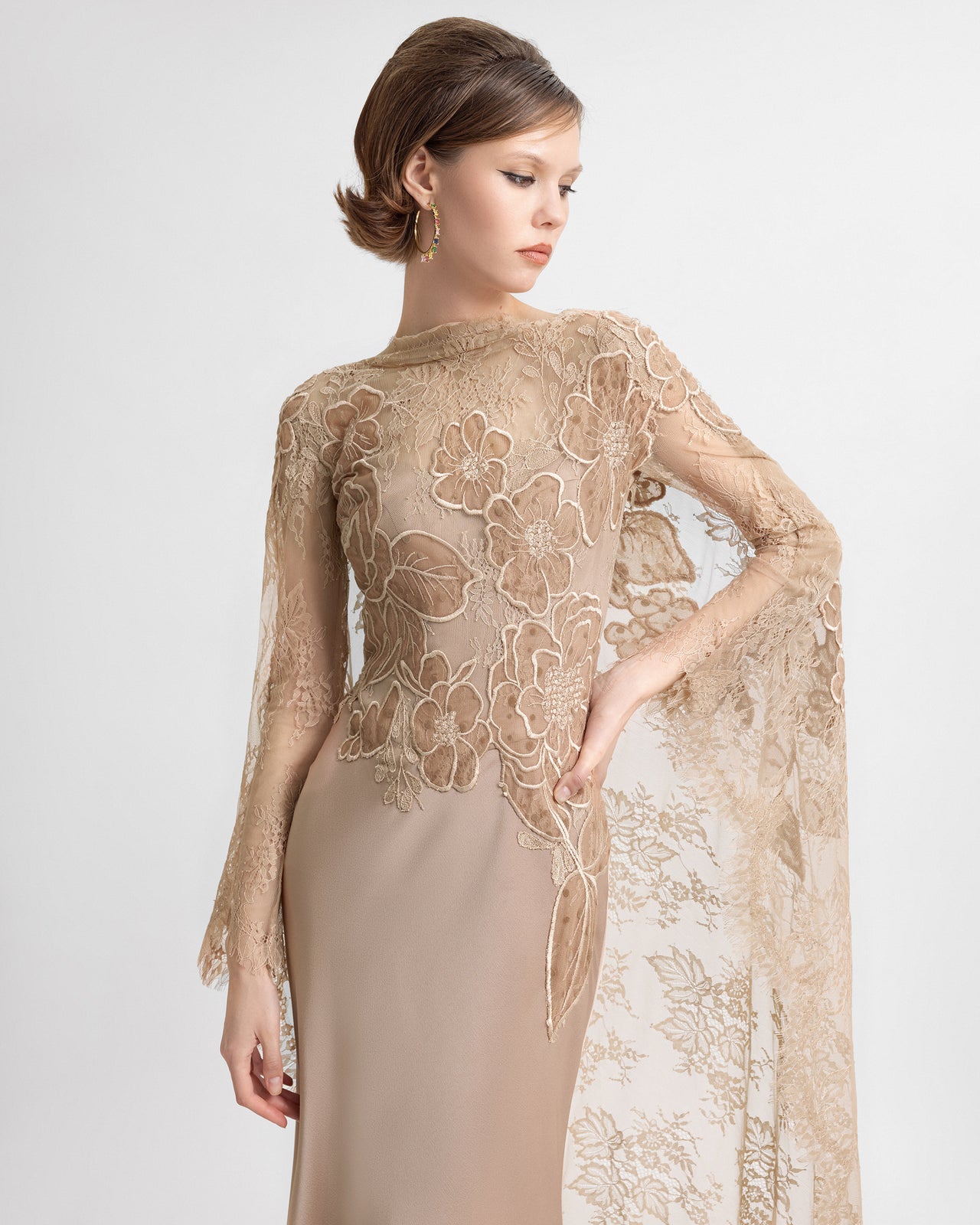 A model walks in a beige floor-length corset gown with a sheer embroidered lace cape. The camera captures the dress’s flowing silhouette, floral lace details, and low scoop back as the model moves gracefully.