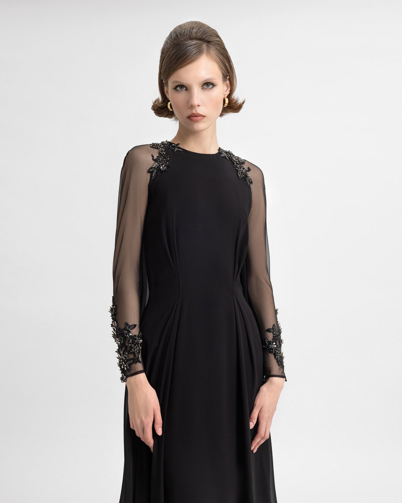 Video of a model wearing a black crepe georgette gown with sheer beaded sleeves and a flowing back cape. The model moves to showcase the shimmer of the embellishments, the lightweight drape, and the gown’s 120cm length from waist to hemline.