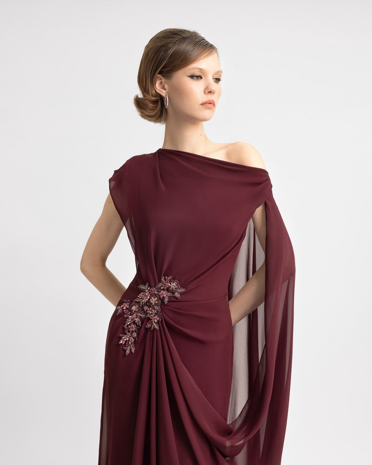 Model walks in a burgundy asymmetrical gown with beaded waist and a flowing cape sleeve.