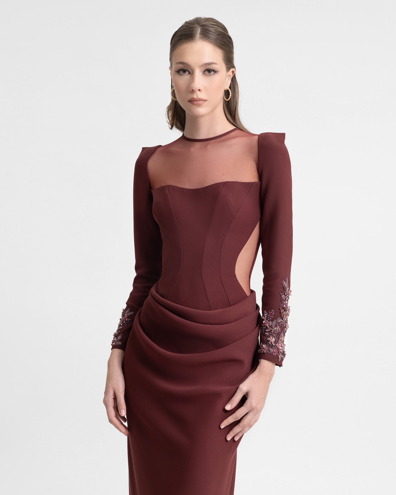 Model walks in a burgundy corset gown with sheer neckline, beaded cuffs, and draped waist detailing. The video captures the dress’s fitted silhouette, sheer buttoned back, and soft train in motion.