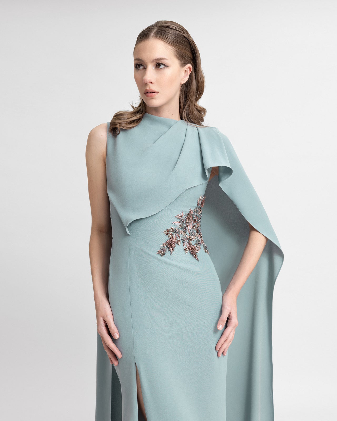 Video of a model wearing a light blue asymmetrical crepe gown with a draped cape, rose gold beading at the waist, and a high side slit, capturing the gown’s graceful flow and sculptural details in motion.