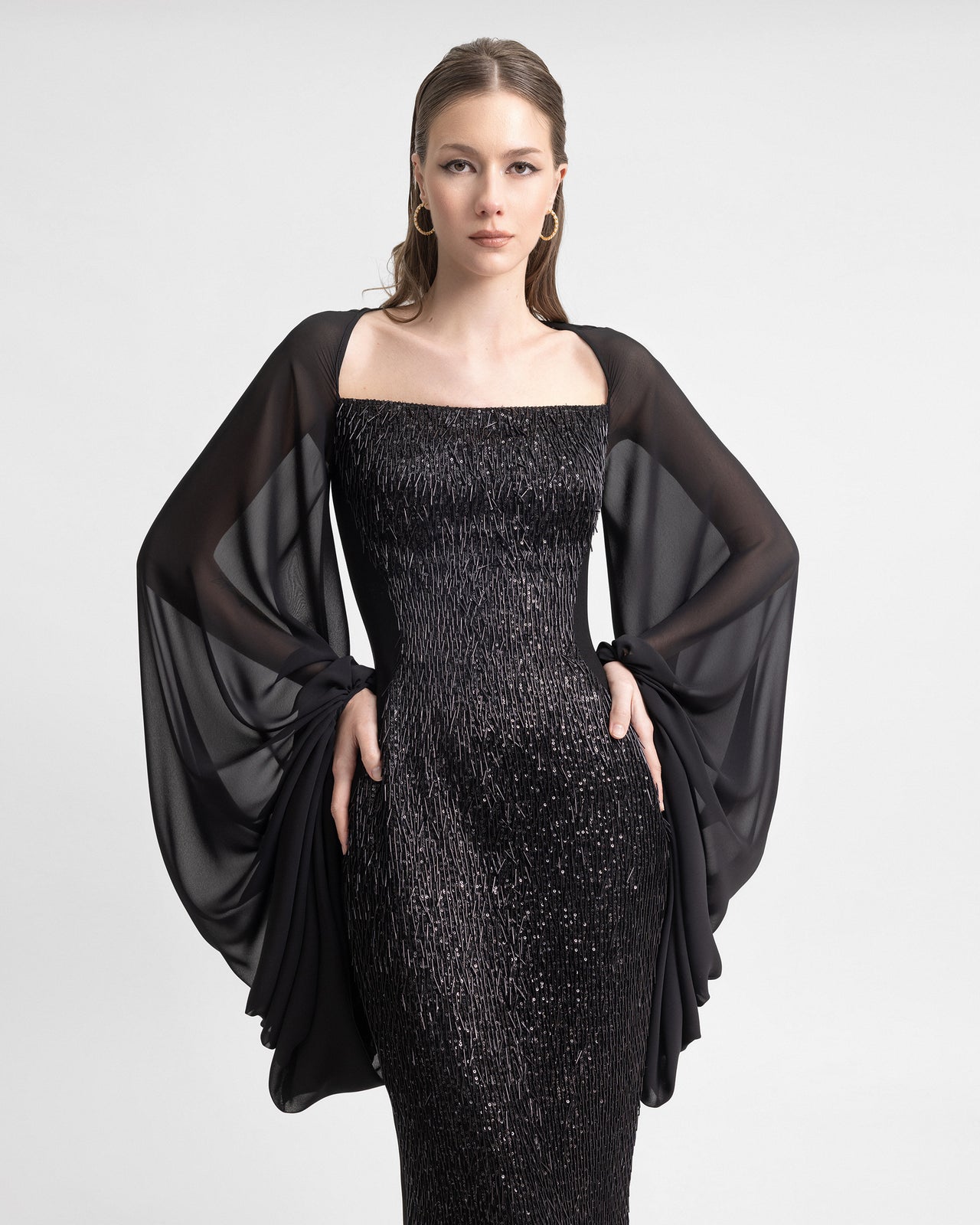 Video of a model wearing a slim-cut black gown with a beaded tassel front and sheer puffed sleeves. The model turns to reveal the back slit and flowing sleeve movement, highlighting the shimmer and sculpted fit of the 120cm-long dress.