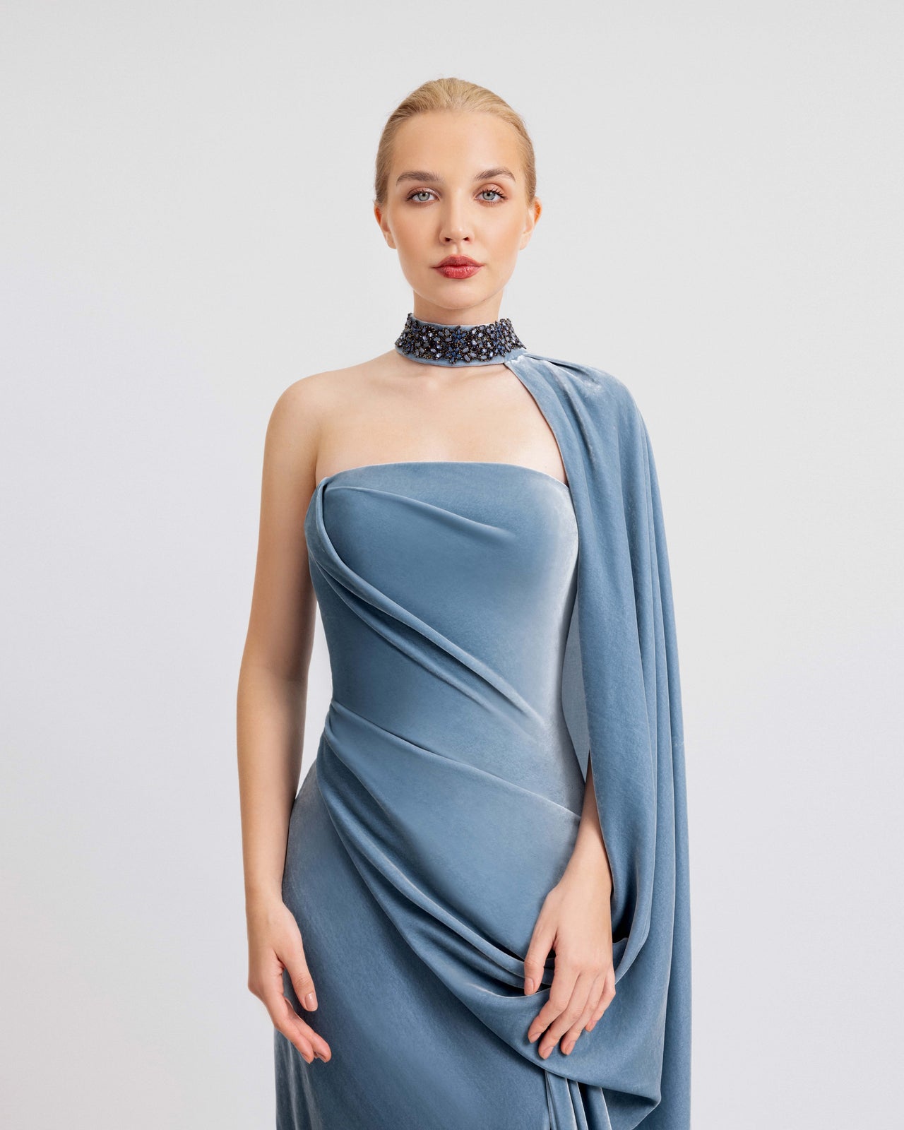 Video of a woman modeling a soft blue velvet evening gown with off-shoulder draping, a high slit, a dramatic train, and a beaded collar, showcasing the dress’s fluid movement and elegant silhouette.