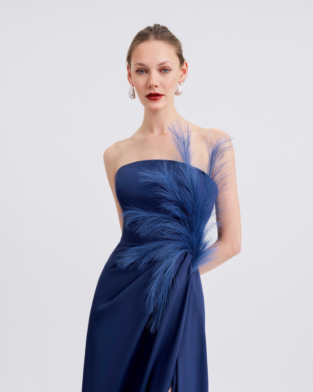 A video for a strapless navy blue evening gown with dramatic feather detailing on the corset and a high slit on the side. The video highlights the gown’s elegant draping, movement, and back view with a fitted silhouette.