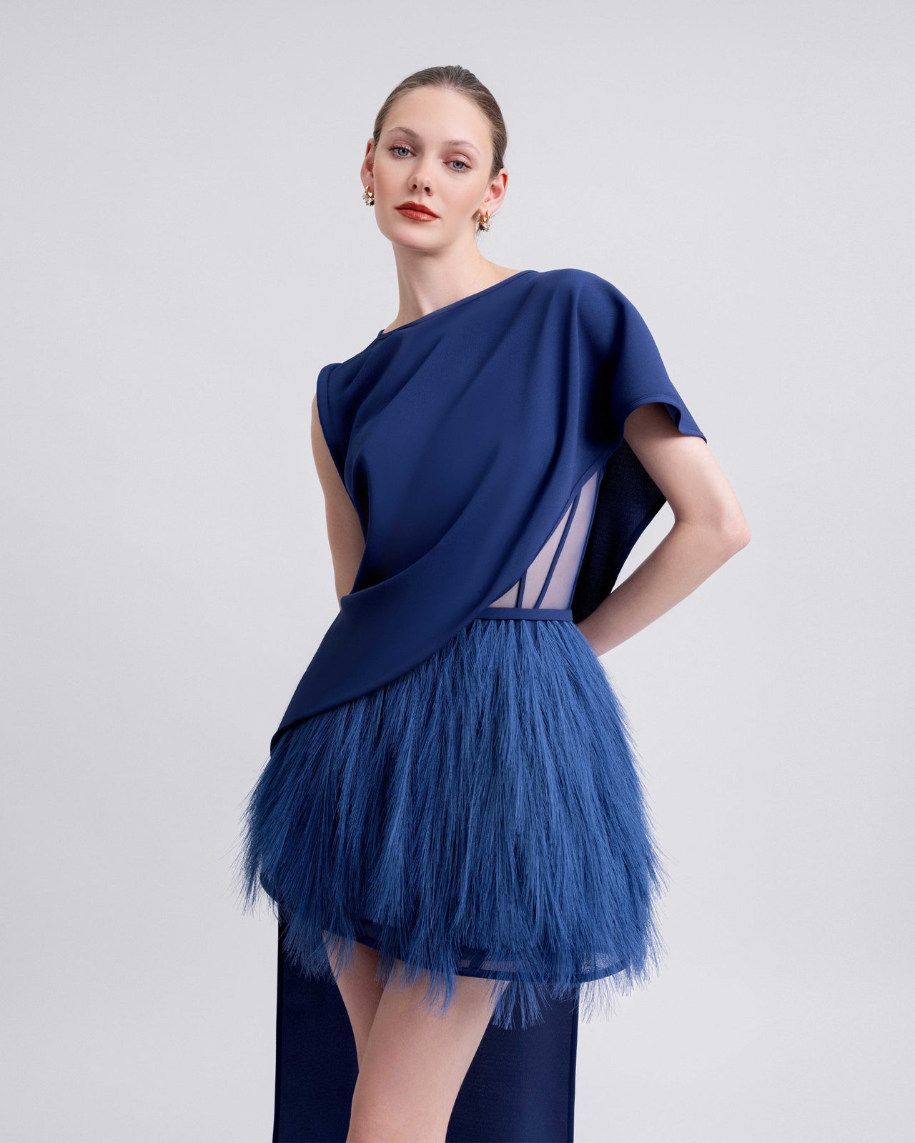 Video showcasing a short blue feathered dress paired with an asymmetrical crepe overlay featuring a long train. The video highlights the contrast in textures, the dynamic movement of the feathers, and the flowing drape from multiple angles.