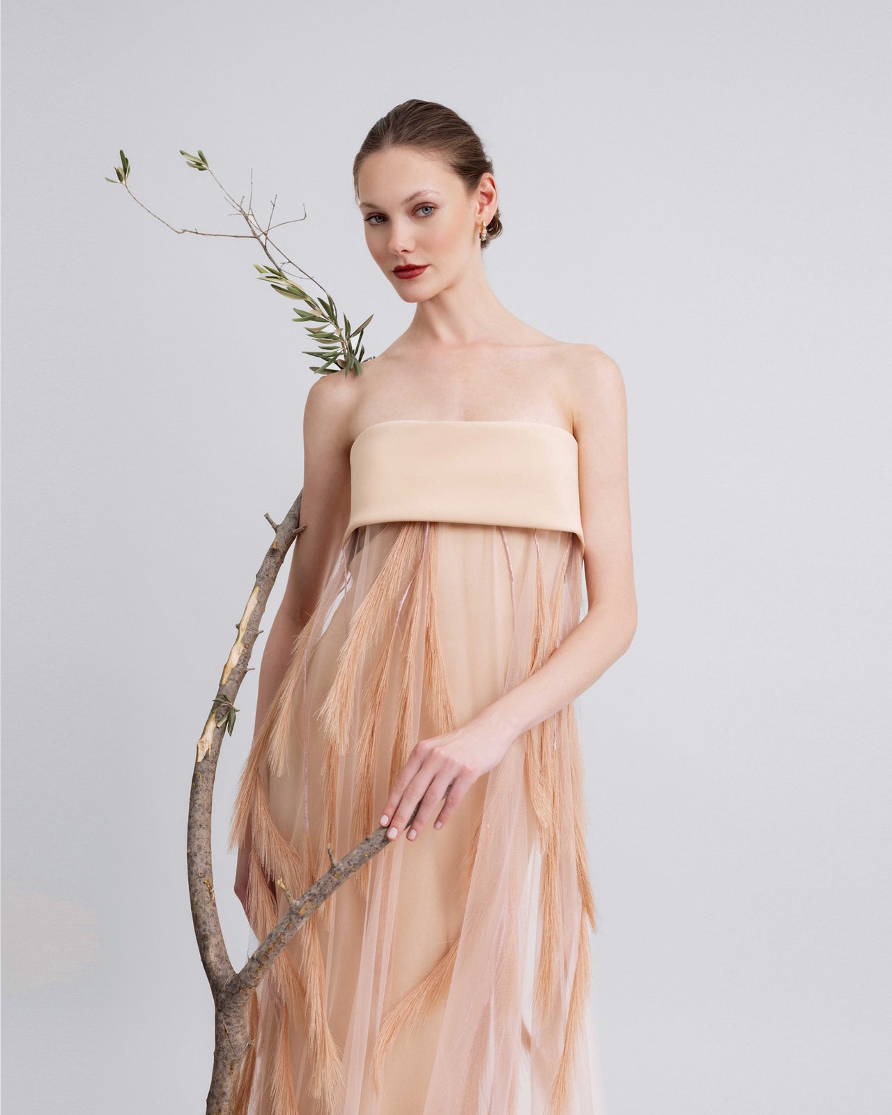Video of a woman modeling a strapless peach gown with a structured crepe bodice and flowing feather-embroidered sheer overlay, highlighting the gown’s soft movement and airy texture.