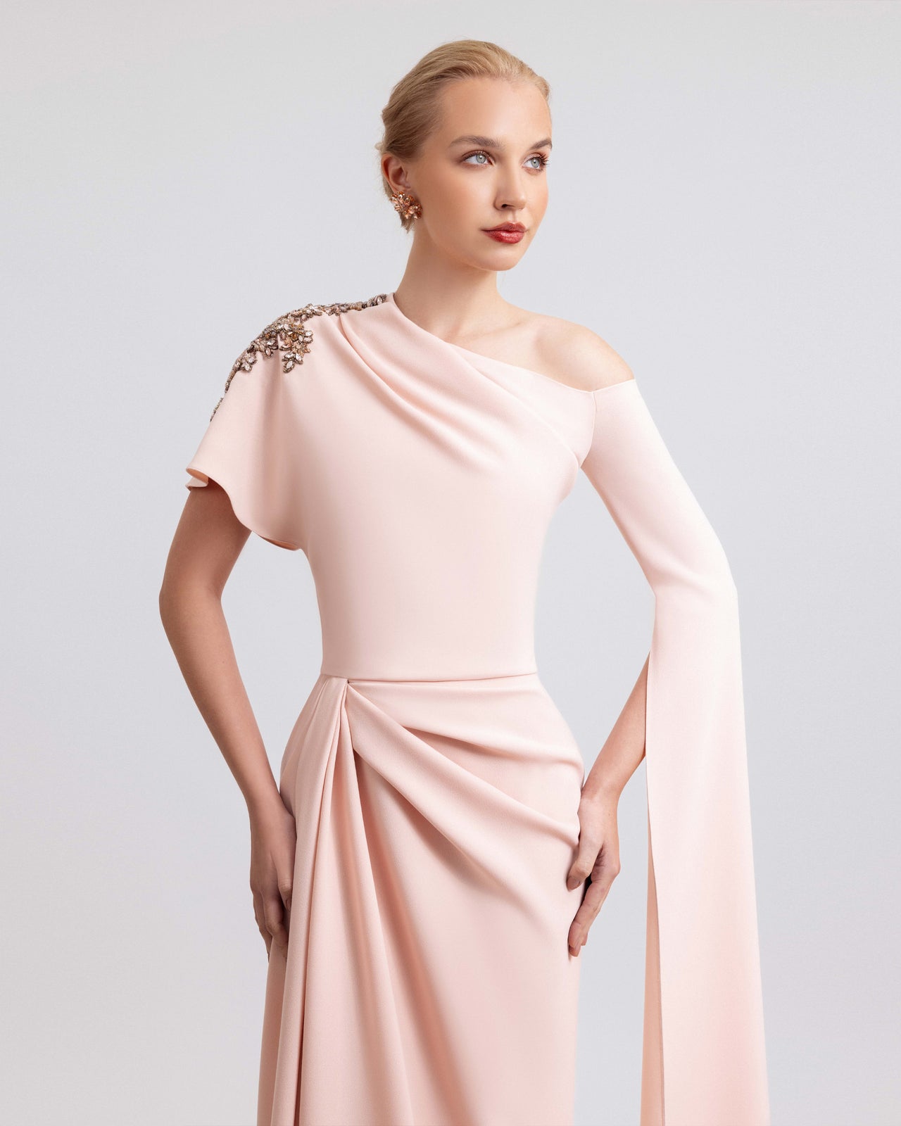 Video of a woman modeling a blush pink crepe gown with an asymmetrical neckline, shoulder beadwork, a floor-length sleeve, and a side slit, highlighting the gown’s elegant drape and fluid movement.