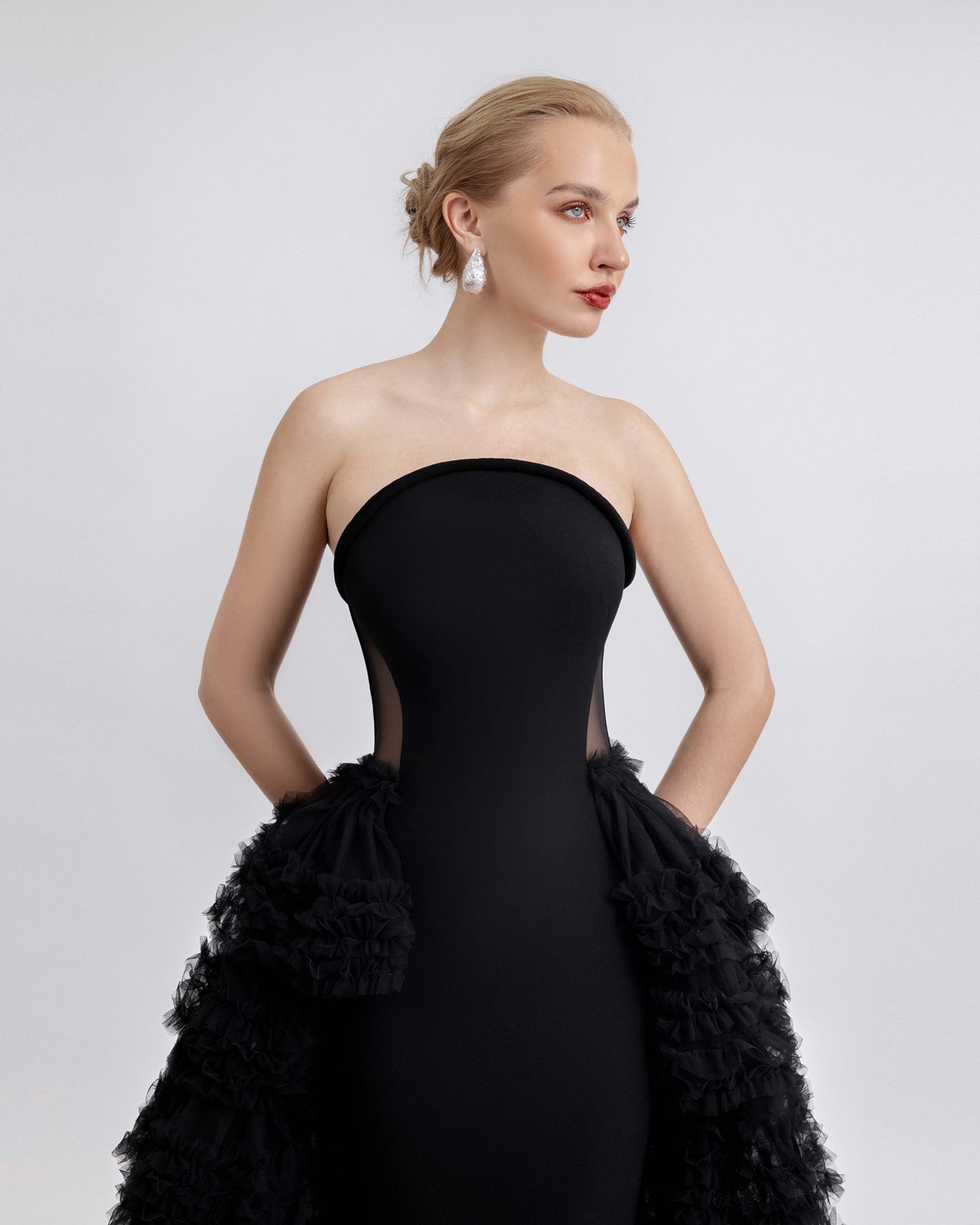 Video of a strapless black gown featuring a structured neckline, sheer mesh side panels, and a fitted crepe silhouette. The gown is complemented by a dramatic tiered tulle overskirt with cascading ruffles. The video captures the model walking and turning to showcase the elegant movement, volume, and intricate details of the dress from all angles.