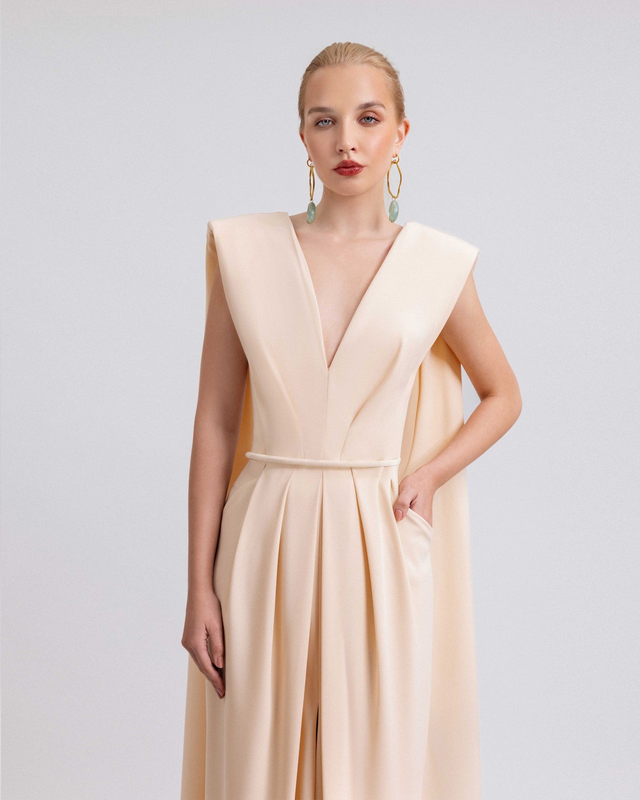 Video of a woman modeling a blush crepe gown with a deep V-neckline, pleated skirt with a center slit, and detachable belt, featuring dramatic shoulder drapes and a structured 120cm silhouette in motion.