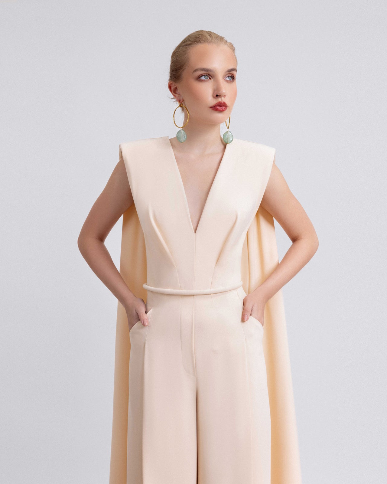 Video of a woman modeling a blush crepe ensemble with a V-neckline top, straight-cut pants, detachable belt, and flowing shoulder drapes, highlighting the structured tailoring and 120cm pant length in motion.