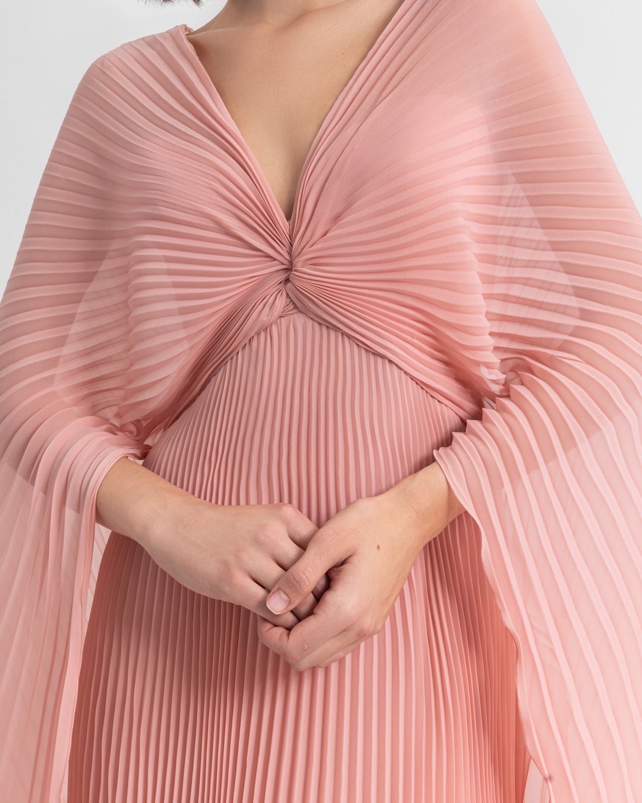 A close-up of a V-neckline fully pleated flared chiffon dress in pink with draping details.